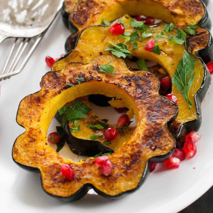 Roasted acorn squash slices garnished with pomegranate seeds and chopped parsley, lined up on a white platter.