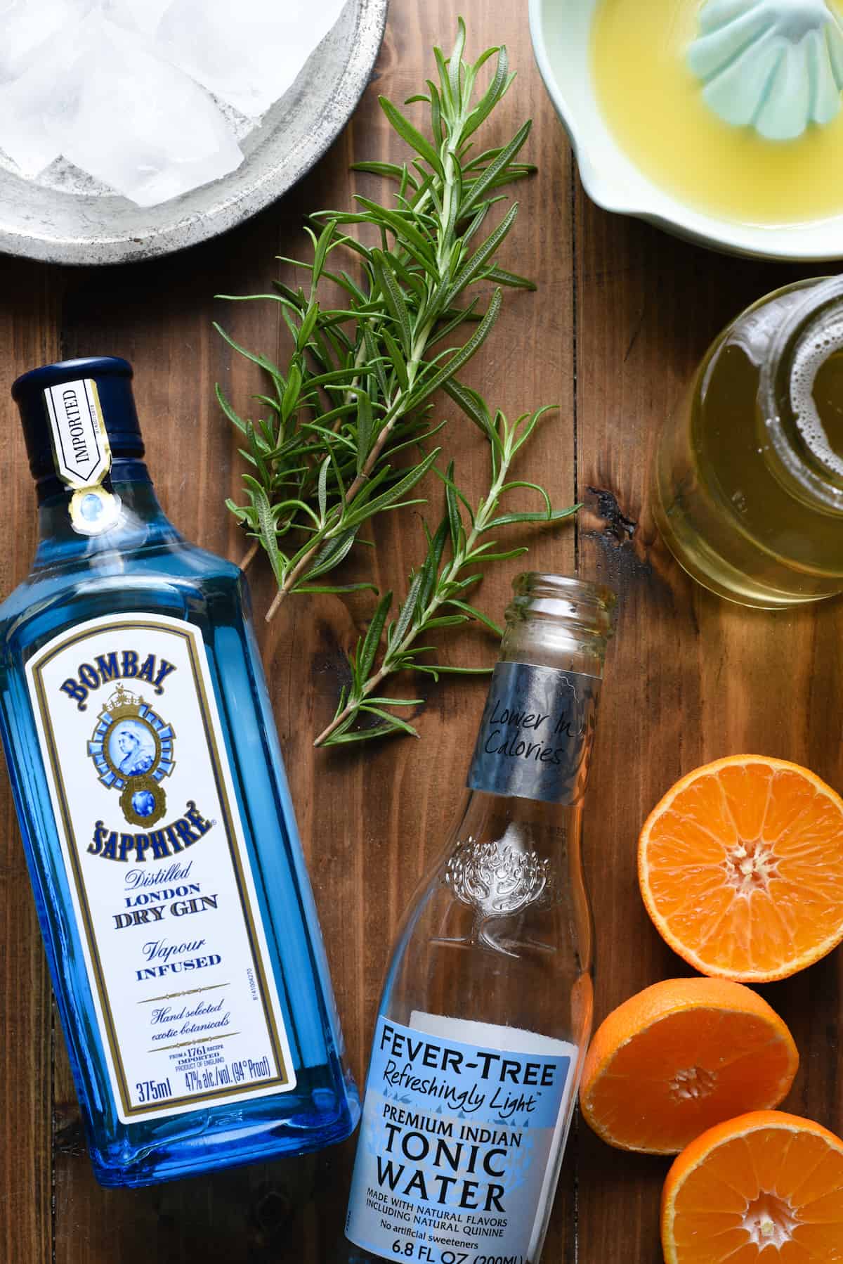 Gin and tonic water bottles, rosemary, cut oranges, ceramic juicer and ice cubes on wooden background.