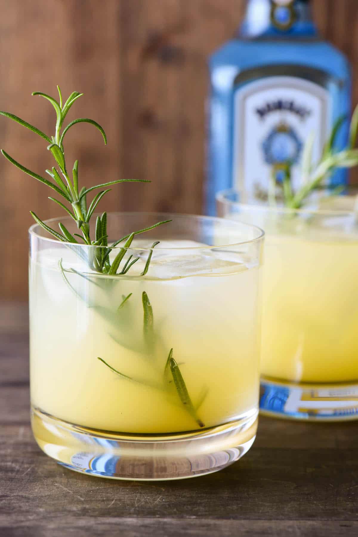 Orange hued rosemary gin cocktail with rosemary sprig garnish, and blue gin bottle in background.