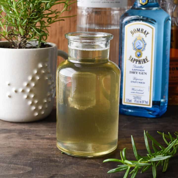 Small bottle of rosemary simple syrup with liquor bottles and rosemary plant in background.