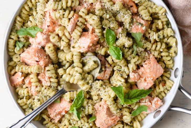 Overhead photo of skillet filled with noodles, a green herb sauce, and chunks of pink fish, with a spoon digging into it.