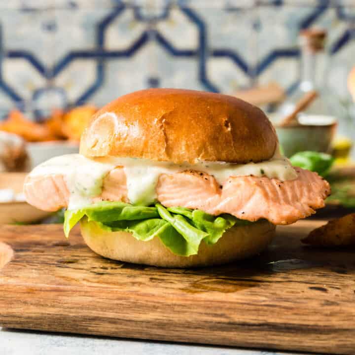 A grilled salmon sandwich with lettuce and mayo on a brioche hamburger bun, on a small cutting board in front of a colorful tile backsplash.