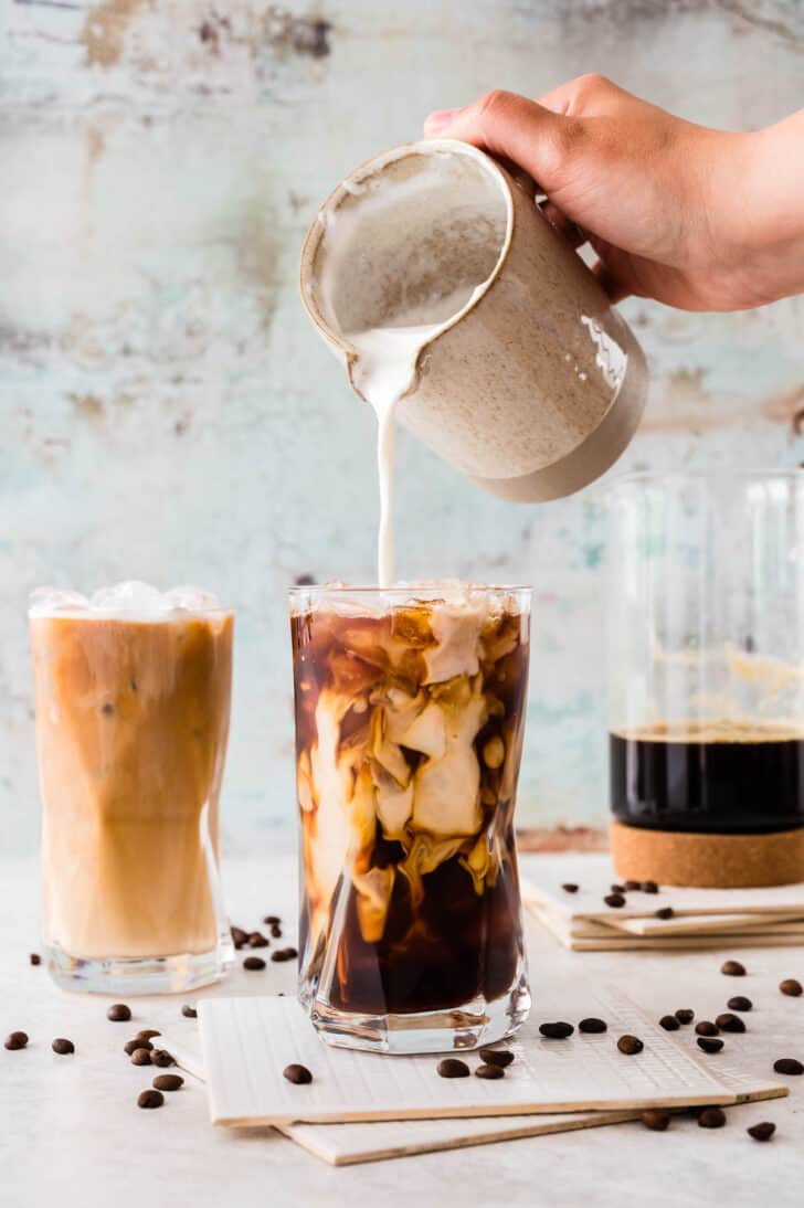 A large glass of iced coffee with cream from a pitcher being poured into it. The scene is garnished with napkins, a pitcher of coffee, and coffee beans.