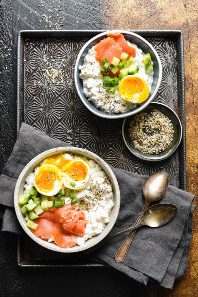 Baking pan with two bowls full of curd cheese topped with soft eggs, smoked salmon, green onions and cucumber, along with spoons and a small dish of everything bagel seasoning.