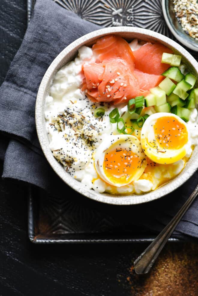 Gray bowl on top of dark gray napkin. Bowl is filled with a savory cottage cheese breakfast, including a soft egg and smoked salmon.