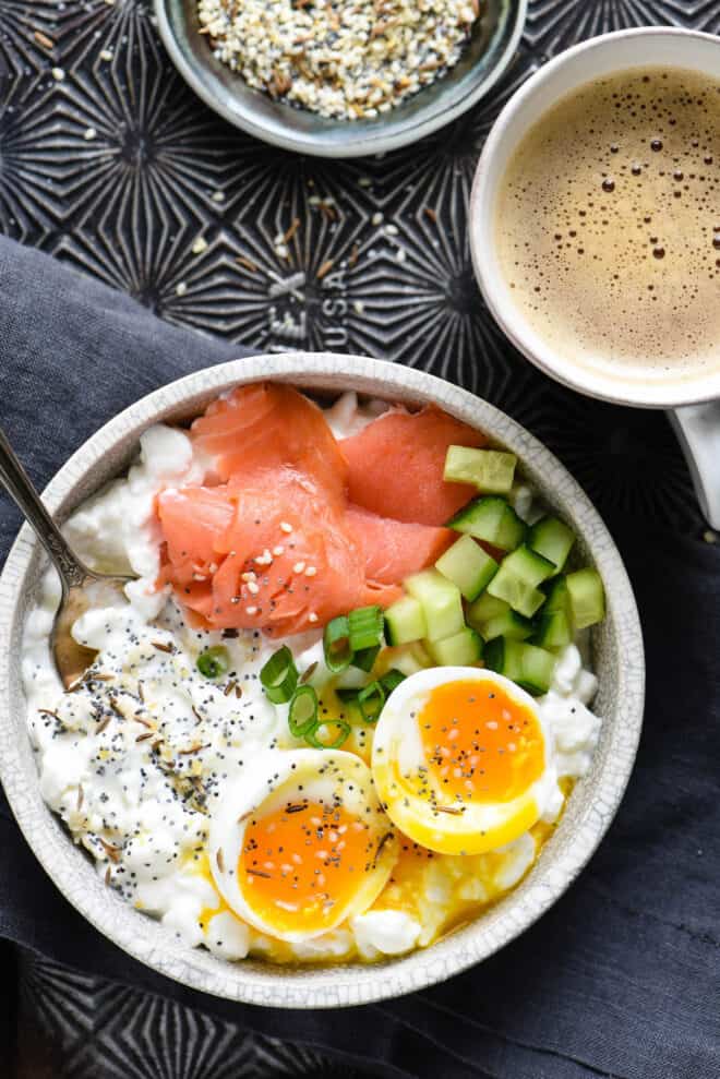 Savory cottage cheese bowl topped with lox, onions, cucumbers and soft egg, alongside mug of coffee and small bowl of seasoning mix.