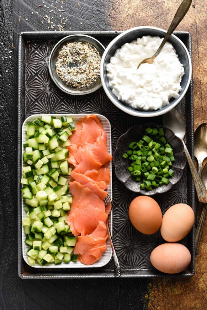 Baking pan with small dishes of everything bagel seasoning, cottage cheese, sliced green onions, brown eggs, chopped cucumber and smoked salmon.