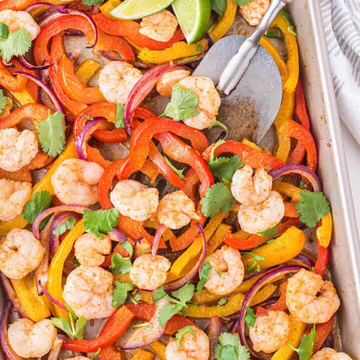 Sheet pan shrimp fajitas sprinkled with cilantro on a light marble surface, with a metal spatula scooping some off the pan.