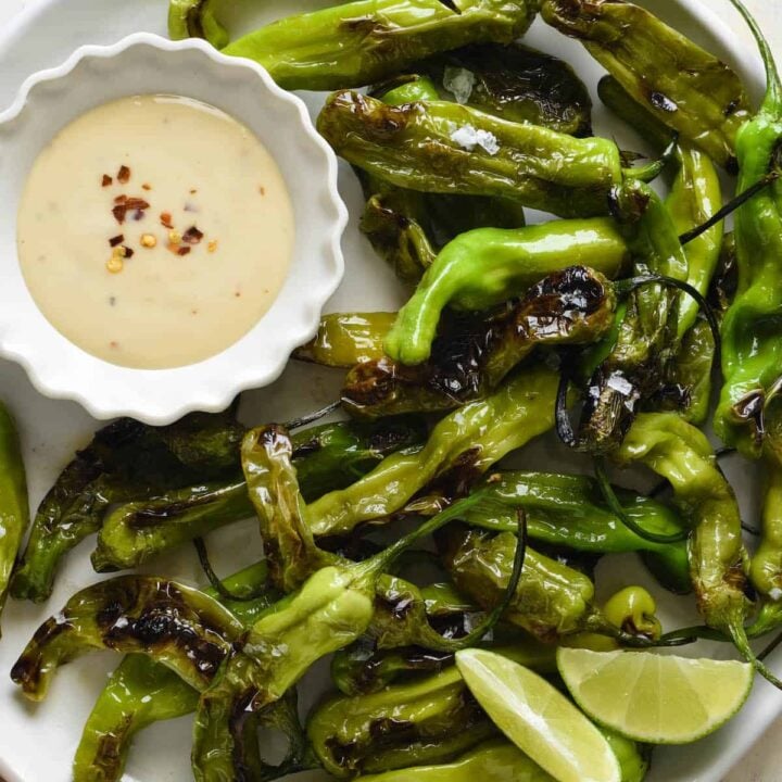 Round white plate filled with blistered shishito peppers and a small fluted white bowl of creamy light orange sauce with red pepper flakes sprinkled on top.