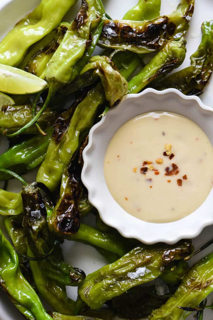 A small white fluted bowl of creamy orange shishito peppers dipping sauce with red pepper flakes, alongside blistered shishito peppers.