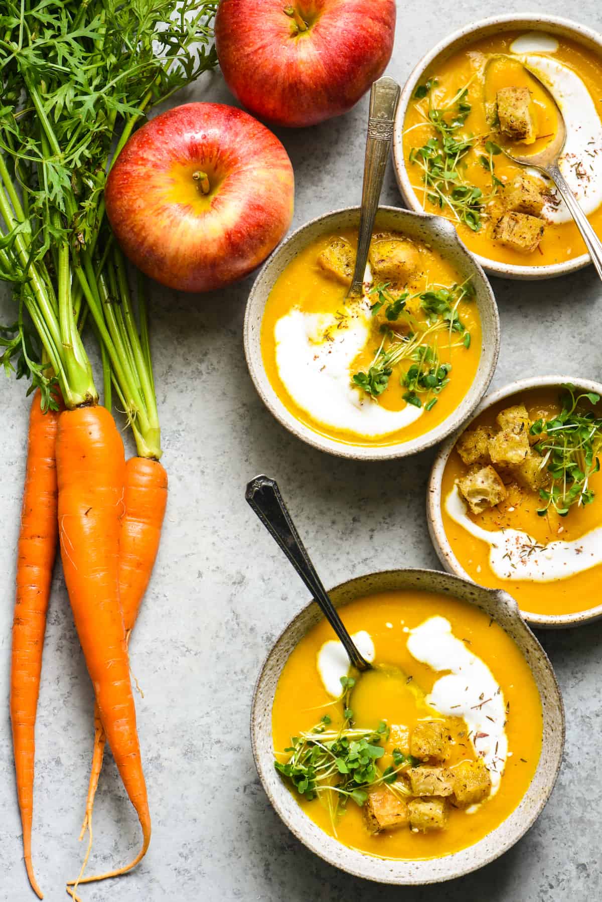 Overhead shot of 4 bowls of pureed carrot soup on gray background, topped with yogurt, herbs and croutons. Apples and carrots on table near soup.