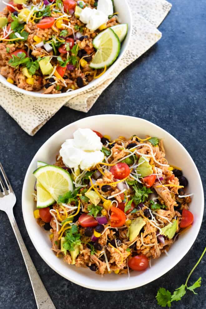 Two white ceramic dishes filled with slow cooker chicken burrito bowl mixture, garnished with sour cream, cilantro and limes.