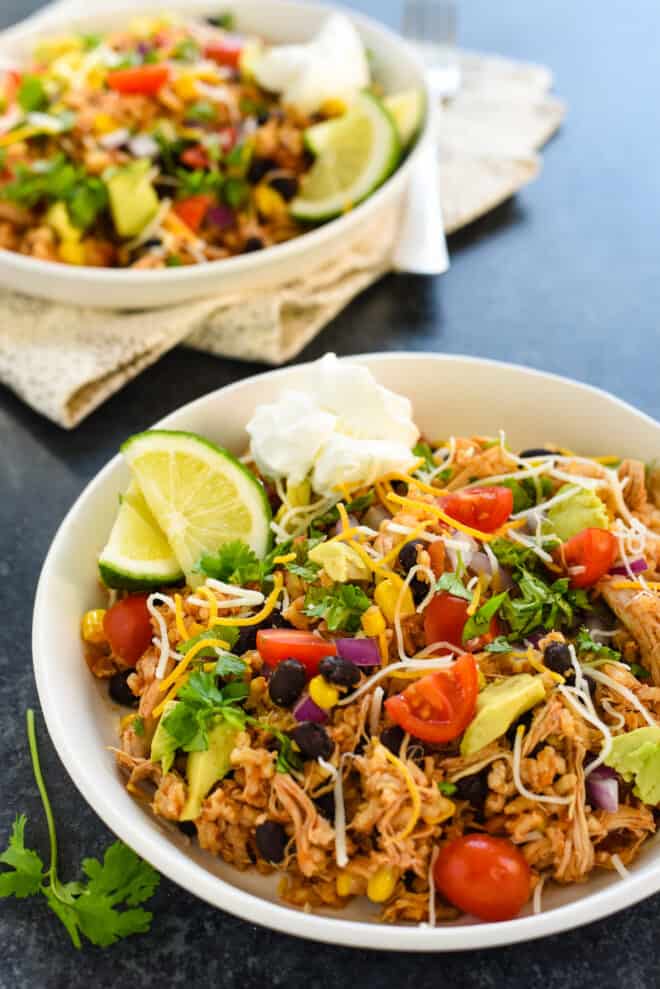 Two shallow bowls filled with a mixture of rice, beans, chicken and vegetables, garnished with sour cream and lime wedges.