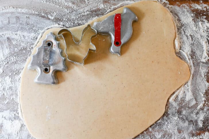 Three cookie cutters (a Christmnas tree, squirrel and chicken shapes) positioned on top of rolled-out cookie dough on a floured surface.