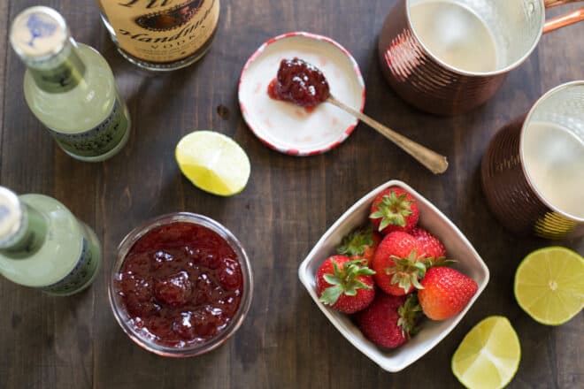 Wooden table with ingredients for strawberry Moscow mules, including strawberries, jam, limes and vodka.