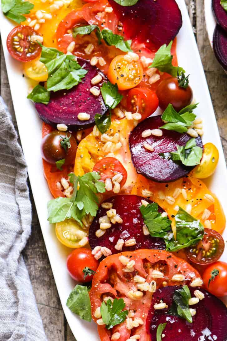 White oblong platter filled with sliced tomatoes, beets, herbs and barley.