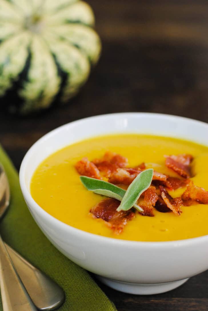 A small white bowl filled with a bright orange pureed soup, topped with crispy bacon pieces and fresh sage leaves.