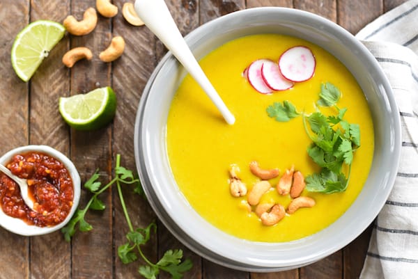 Overhead photo of thai potato soup on wooden tabletop, with cashews, lime wedges, cilantro and hot sauce on table.