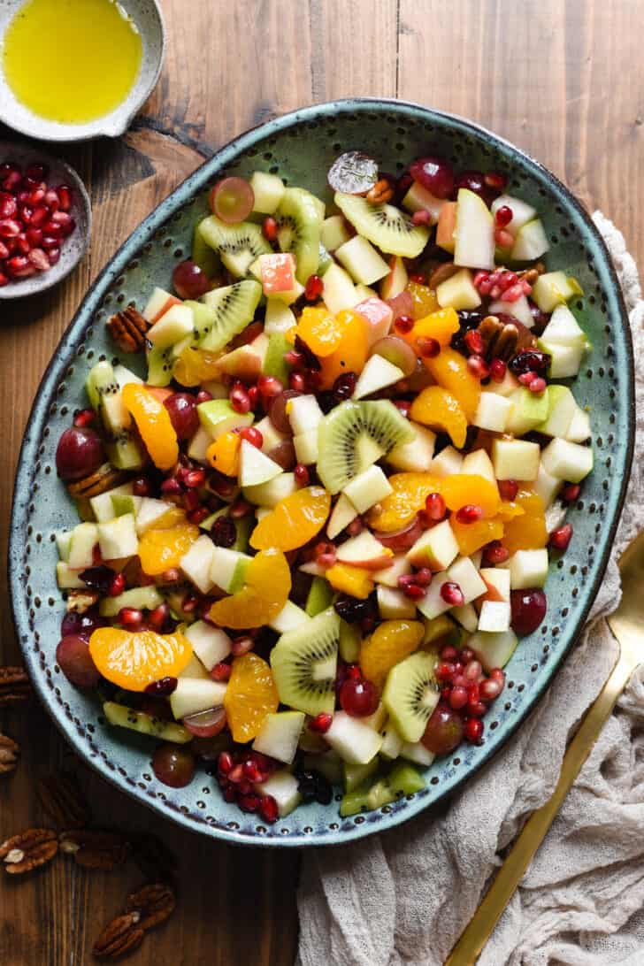 Oval turquoise ceramic bowl filled with Thanksgiving fruit salad made with oranges, grapes, apples, pears, kiwi, pomegranate seeds, dried cranberries and pecans.