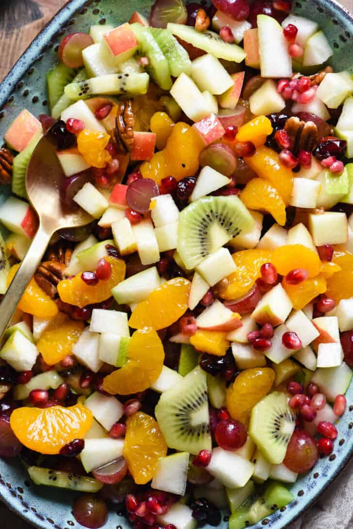 Oval turquoise ceramic bowl filled with chopped oranges, grapes, apples, pears, kiwi, pomegranate seeds, dried cranberries and pecans, with a gold spoon spooning some out.