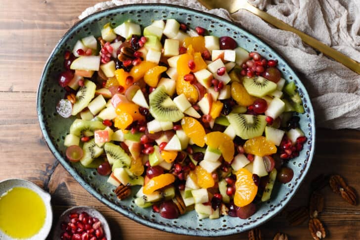 Oval turquoise ceramic bowl with a mixture of chopped oranges, grapes, apples, pears, kiwi, pomegranate seeds, dried cranberries and pecans.