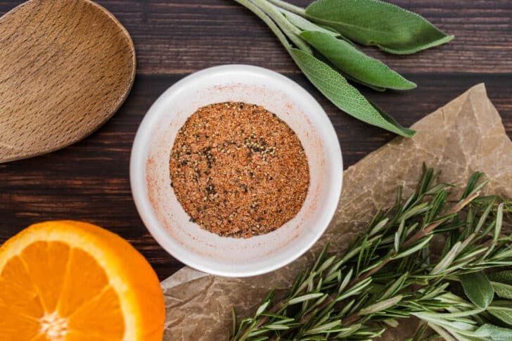 A brown spice rub in a small white bowl, surrounded by fresh herbs.