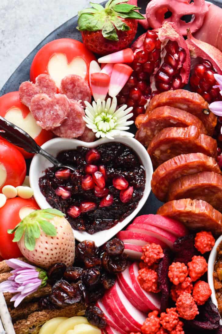 Close up of Valentine's snacks including salmi, strawberries, red jam, pomegranate seeds and Babybel cheese with hearts cut in their wax wrappings.