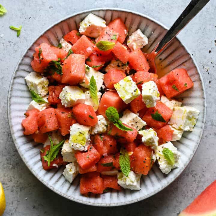 A textured gray bowl filled with watermelon feta mint salad, with a spoon digging into it.