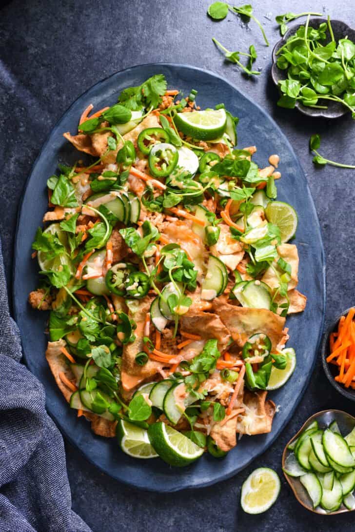 A large slate blue platter filled with wonton nachos, with small bowls of garnishes like herbs and pickled veggies alongside.