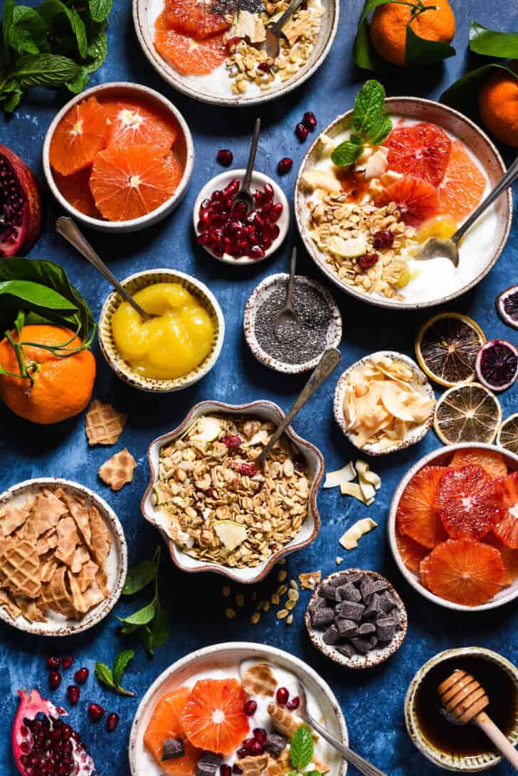 A yogurt bar set up on a blue background, with bowls of toppings including granola, waffle cone chunks, lemon curd, fruit, coconut, oranges and chocolate chunks.